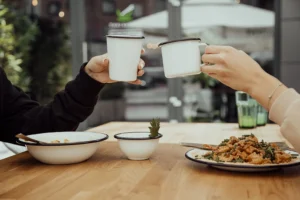 The photo captures an outdoor scene outside a restaurant. On a table, you can see white enamelware, including a deep bowl on the left, a small bowl in the middle, and a shallow plate with an oriental dish on the right. Above the table, two hands are visible, each holding a white enamel mug.