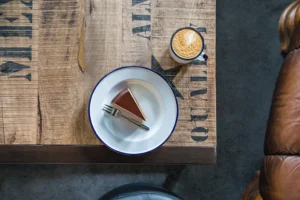 The photo displays a wooden table featuring a white enamel plate with a triangular piece of chocolate cake. Adjacent to it, there is a white enamel mug filled with coffee. Beside the table, a section of a leather armchair is also visible.