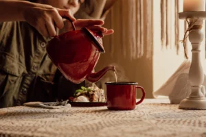 In the center of the photo, there is a red enamel mug and a hand holding a red enamel jug, pouring tea into the mug. Behind the mug, there is a red enamel plate with a piece of cake and ice cream. The person pouring tea is dressed in a green shirt. On the right side of the table, there is a tall white candlestick.