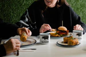 The photo features a white table with two people dressed in black seated at it. On the left side, only hands holding corn are visible, while in front, there is a smiling woman. On the table, you can see three white enamel plates, one of which holds a tall burger, another contains boiled corn. Next to the plates, there are two white enamel mugs featuring a black Butcher logo.