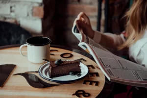 The photo displays a wooden table featuring the word "COFFEE." On the table, there is a white enamel plate and an enamel white mug. The plate holds a chocolate cake with blueberries and a fork. A woman is seated at the table, holding a newspaper in her hands. Behind the table, there is a brick wall.