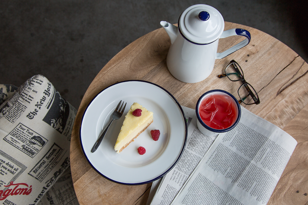 The photo depicts a round wooden table adorned with a white enameled plate holding cheesecake and raspberries, a white enamel mug, and a white enamel coffee pot. On the table, you can also see a pink drink with ice cubes, a newspaper, and a pair of glasses placed next to the mug. Adjacent to the table, there is an armchair with fabric upholstery.