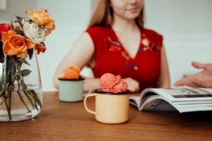 In the photo, in the foreground, there is a wooden table with a glass vase filled with roses and two enamel mugs containing ice cream. The first mug has yellow ice cream, while the one behind it holds orange ice cream. In the background, a woman with long hair, dressed in red, is seated at a table.