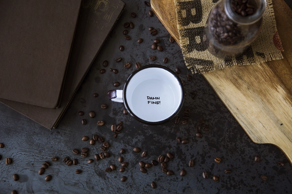 A top-down view reveals the focal point – a white enamel mug proudly bearing the words 'DAMN FINE!' on its bottom. Scattered coffee beans surround the mug. To the left, two menu cards are elegantly framed in brown, while on the right, a wooden cutting board rests beneath a canvas piece and a glass bottle brimming with coffee beans.