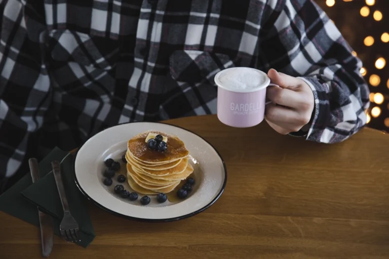 The photo features a wooden table where a person in a black and white checkered shirt sits, holding a pink enamel Gardelli-inscribed mug. On the table, there's a white enamel plate with pancakes and blueberries, accompanied by a knife and fork.