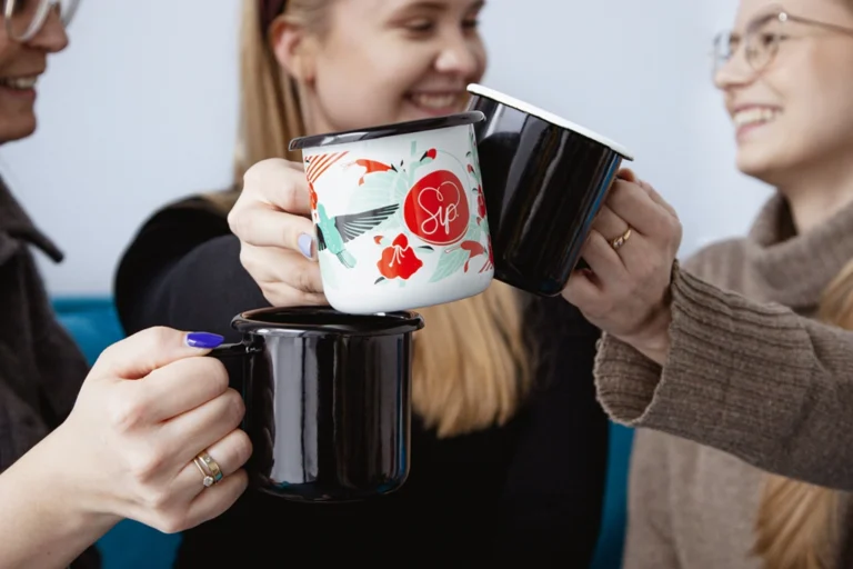 In the foreground of the photo, you'll find three enamel mugs: two black and one white with colorful patterns. These mugs are joyfully held by three laughing women in the background of the photo.