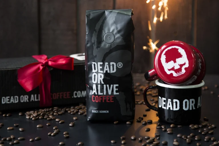 In the central part of the photo, there is a large black package of coffee with the inscription 'DEAD OR ALIVE COFFEE, GANZE BOHNEN.' On the right side, there are two enamel mugs: a black one at the bottom with the inscription 'DEAD OR ALIVE' and a red one with a graphic featuring a white skull on the bottom. On the left side, behind the coffee, there is a black cardboard box tied with a red ribbon.